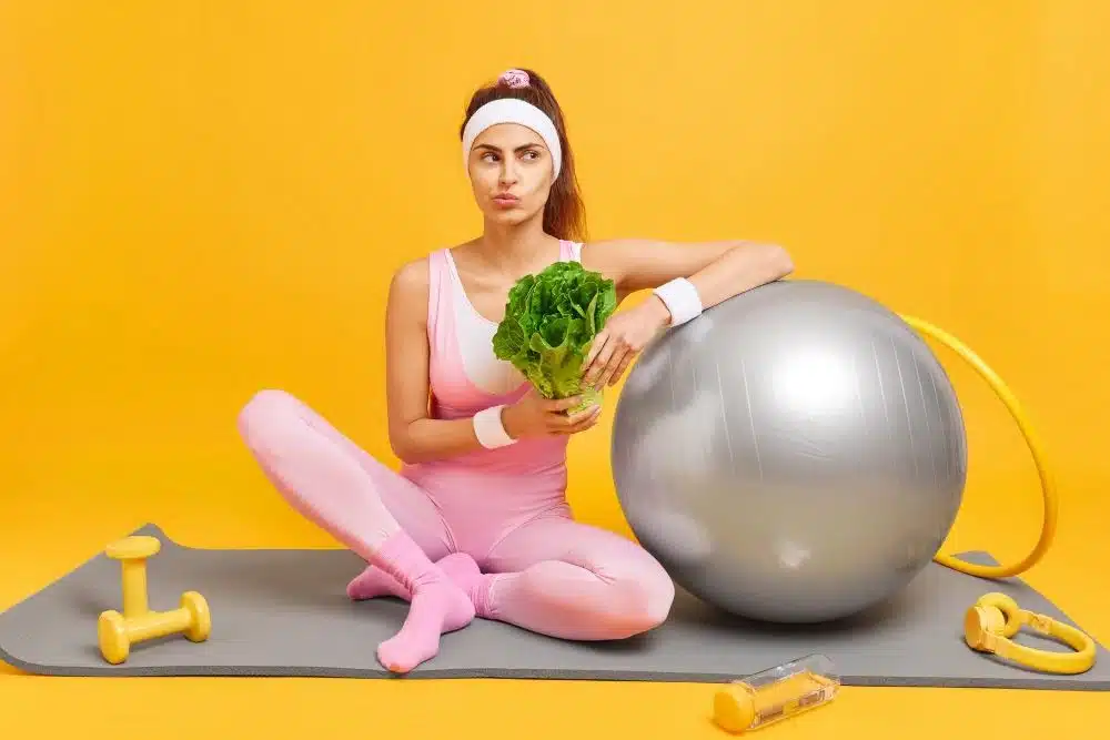 Woman sitting on mat holding green veg thinking which diet and exercise for body type