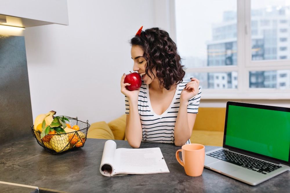 woman holding apple sitting in a modern apartment with fruits in a basket as nutrition hacks, coffee mug and laptop on the table